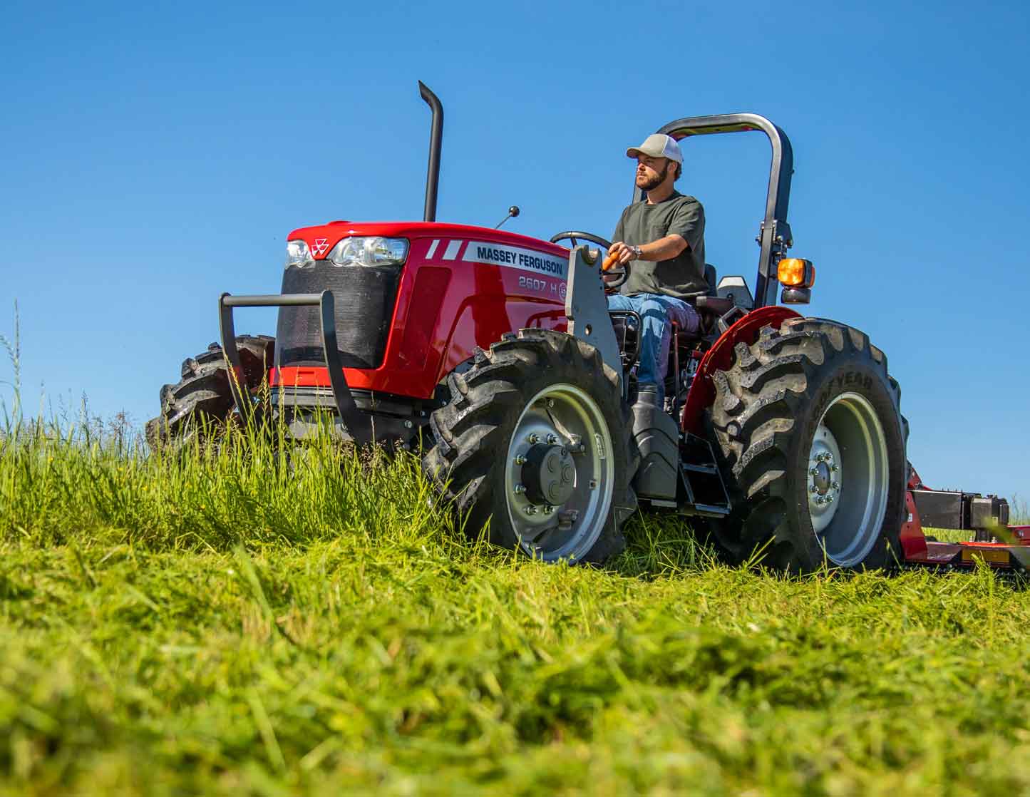 A red Massey Ferguson 2600H tractor driving through a field of freshly cut grass on a sunny day. A man wearing a cap operates the tractor, with a clear blue sky in the background.