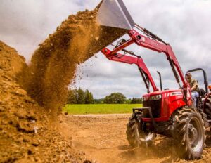 A red Massey Ferguson 2600H tractor lifting and dumping a load of dirt on a sunny day, with green grass and trees in the background. The tractor is equipped with a front loader attachment.