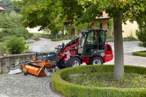 Weidemann 1280 compact loader with front-mounted sweeper attachment cleaning cobblestone paths in a landscaped estate setting.
