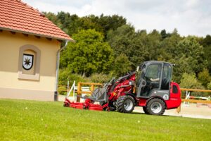 Weidemann 1280 wheel loader equipped with a mower attachment cutting grass near an equestrian facility.