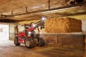 Weidemann 1345T compact telehandler lifting a square hay bale in a low-clearance barn interior