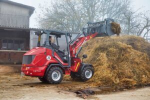 Weidemann 1390 articulated wheel loader moving compost or hay with front loader bucket near a barn.