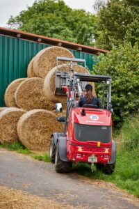 Weidemann 1390 compact wheel loader stacking round hay bales with bale fork attachment on a farm.