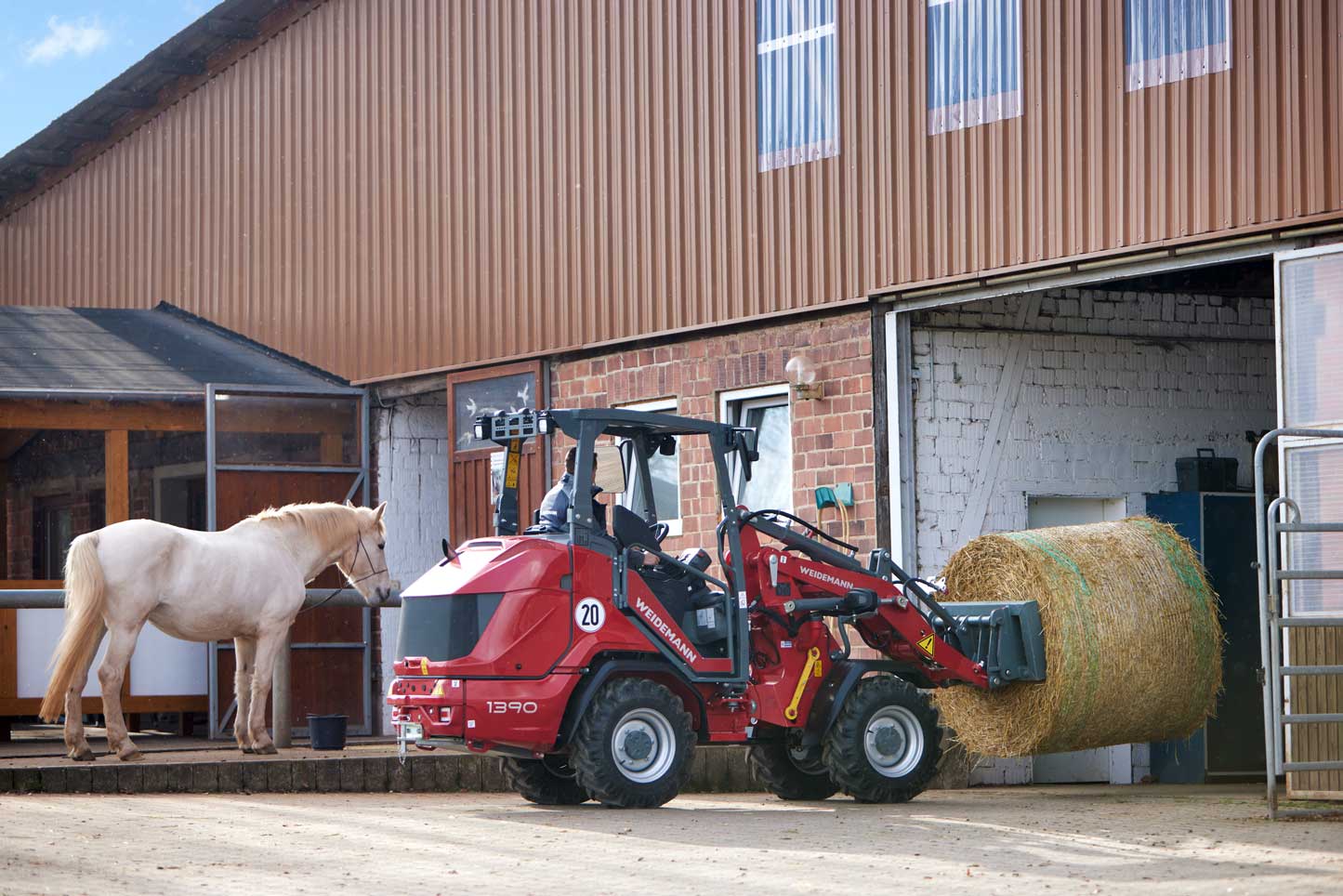 Weidemann 1390 farm loader transporting hay bale into a stable near a horse barn.