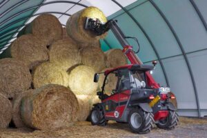 Weidemann T4512 compact telescopic loader stacking large round hay bales inside an arched agricultural shelter