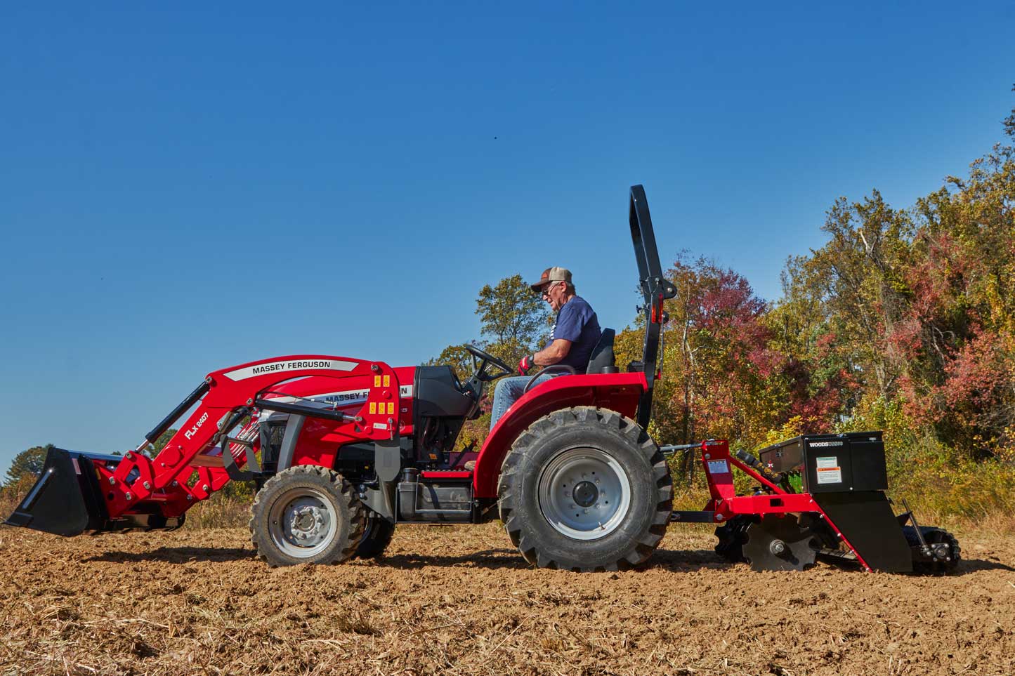 A man operates a red Massey Ferguson 1E Series compact tractor equipped with a front loader and rear-mounted seeder attachment, working in a freshly tilled field under a clear blue sky with colourful autumn trees in the background.
