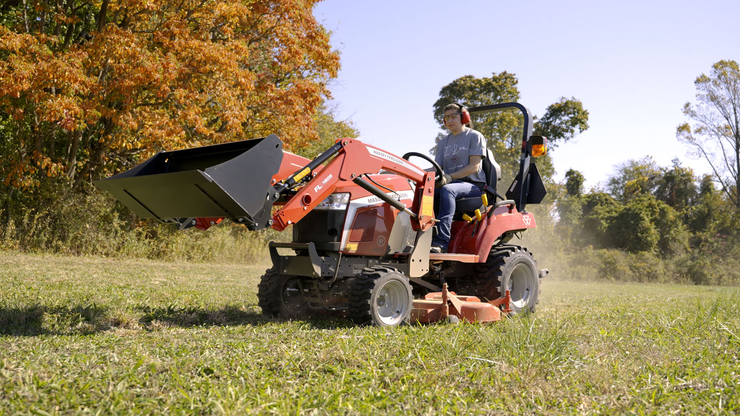 A person operating a red Massey Ferguson 1GC Series compact tractor with a front loader attachment in a grassy field, surrounded by trees with autumn-colored leaves under a clear blue sky.