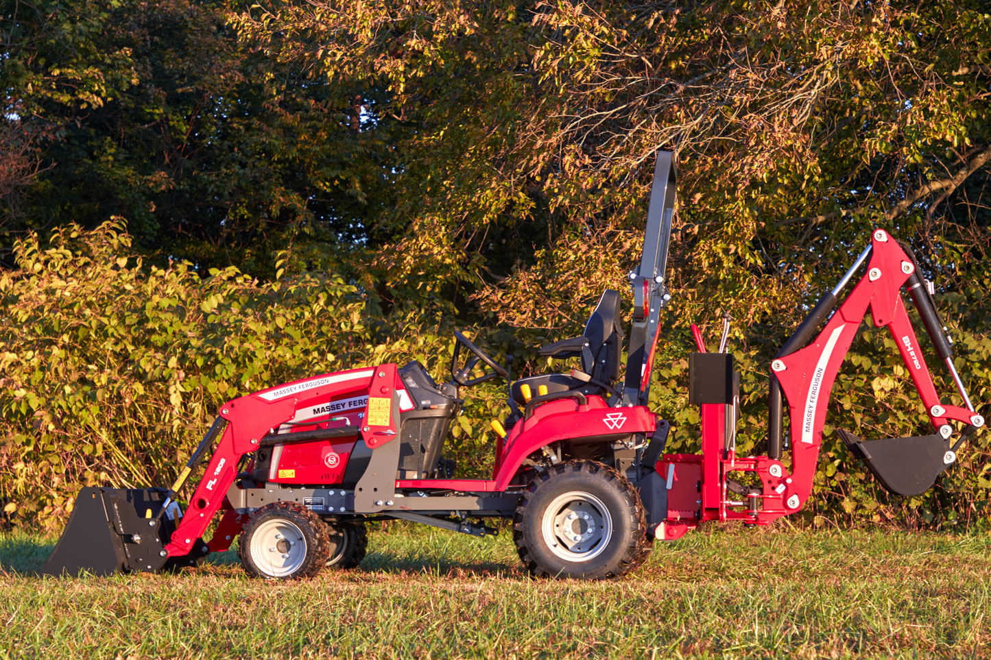 A side profile of a red Massey Ferguson 1GC Series compact tractor equipped with a front loader and a backhoe attachment, parked on grassy terrain with dense green foliage in the background.