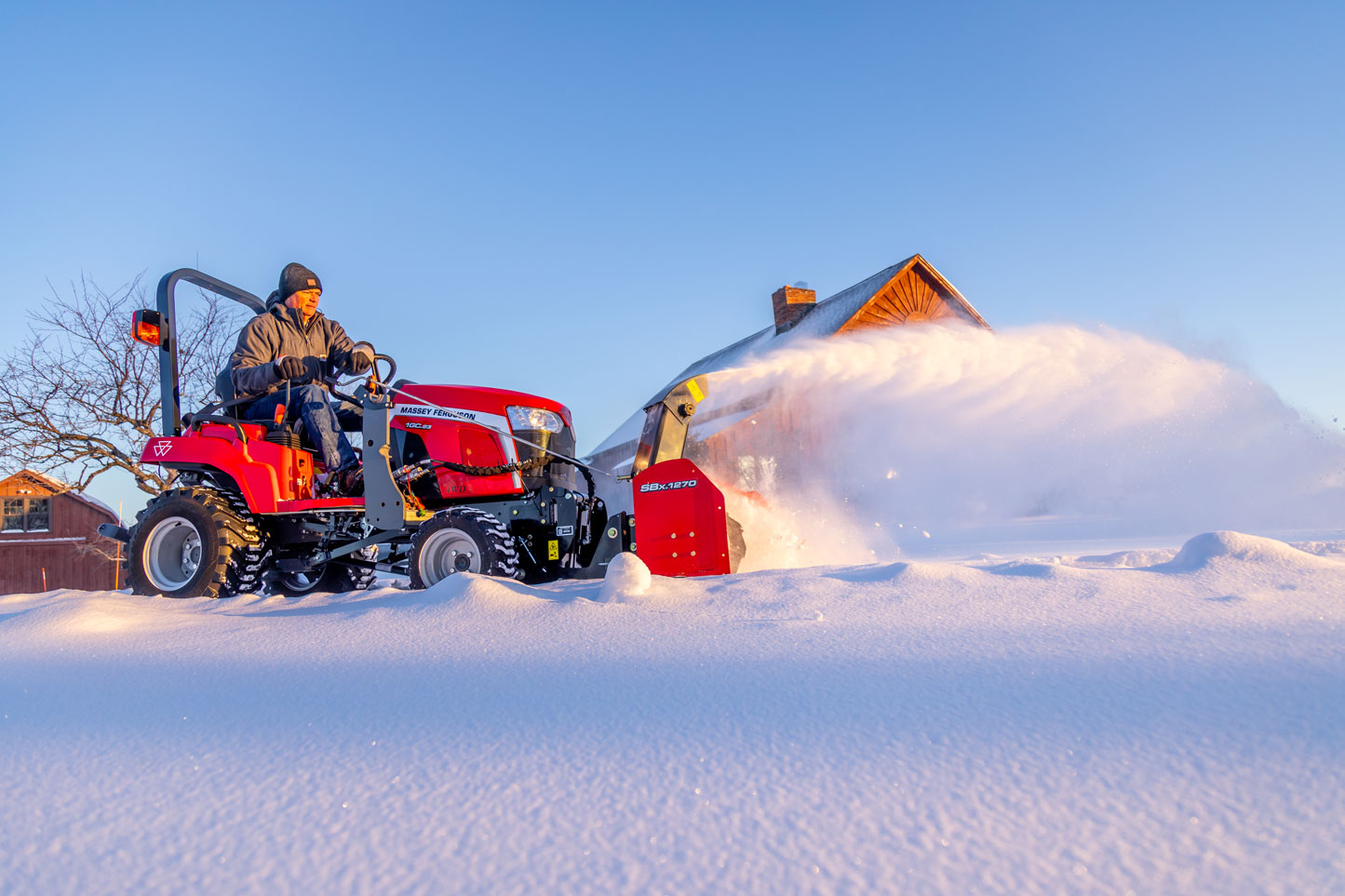 A person operating a red Massey Ferguson 1GC Series compact tractor with a snowblower attachment, clearing snow in a sunlit snowy landscape with a wooden house and bare trees in the background
