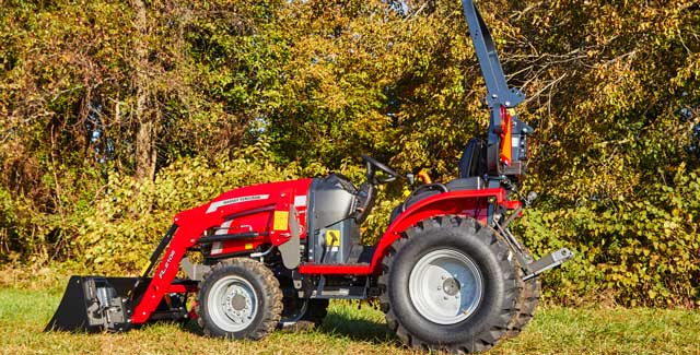 Side profile of a red Massey Ferguson 1M Series compact tractor with front loader, parked on grass in front of dense autumn foliage.