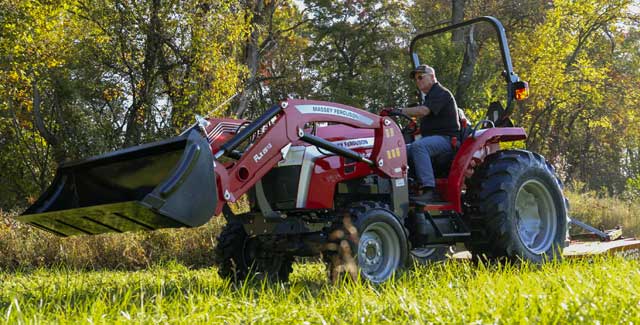A person driving a red Massey Ferguson 2E compact tractor with a front loader and rear mower attachment, mowing grass in a sunlit field surrounded by trees.