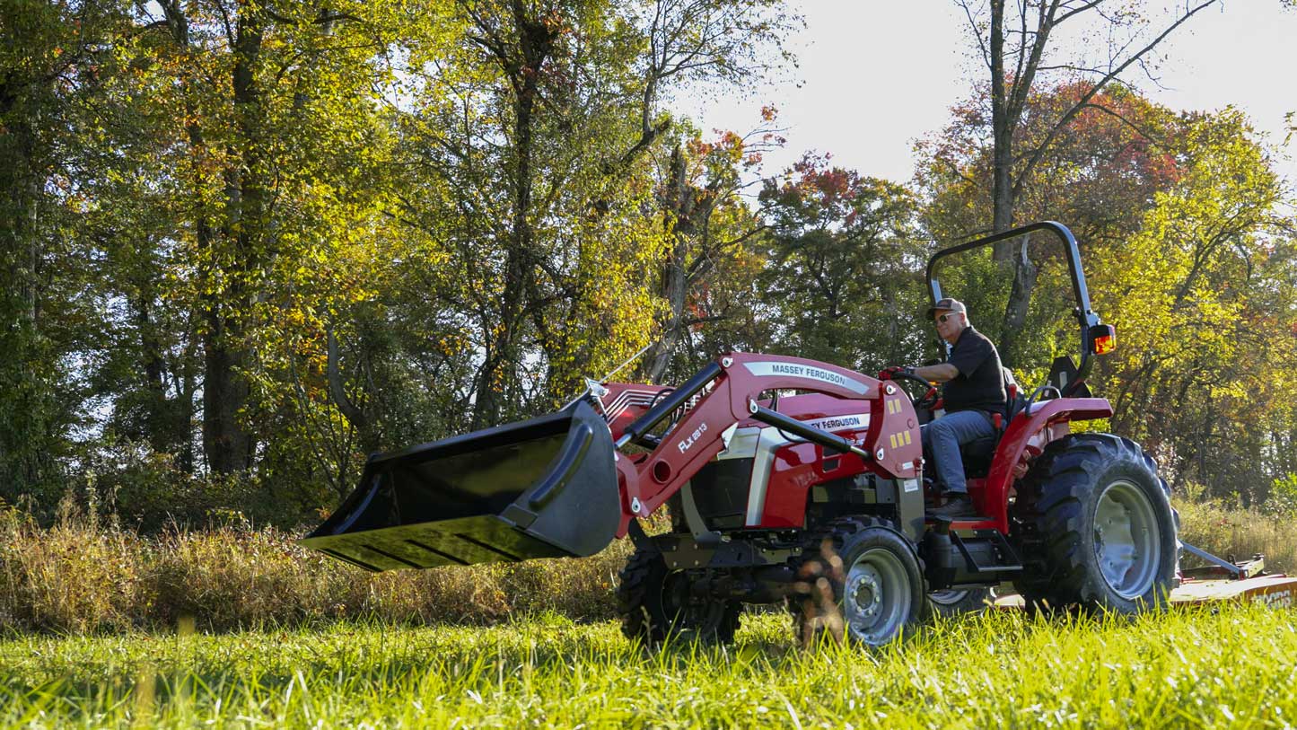 A person driving a red Massey Ferguson 2E compact tractor with a front loader and rear mower attachment, mowing grass in a sunlit field surrounded by trees.