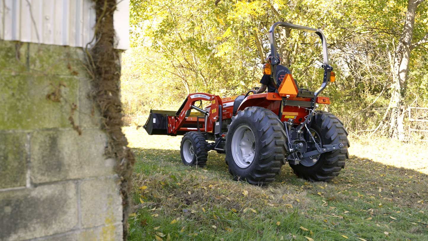 A person operating a red Massey Ferguson 2E compact tractor with a front loader attachment, working near a building in a wooded, leaf-strewn area.