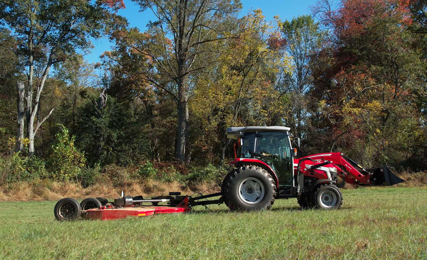 A red Massey Ferguson 2M tractor pulling a large rotary mower across a grassy field bordered by tall trees in early autumn.