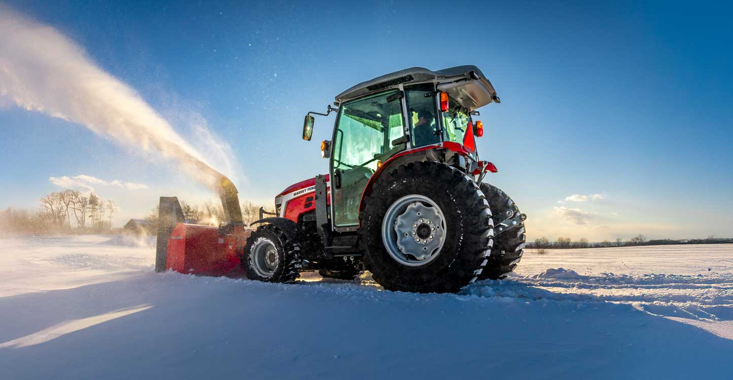 A red Massey Ferguson 2M tractor equipped with a snow blower attachment clearing snow in a wide-open field under a clear blue sky.