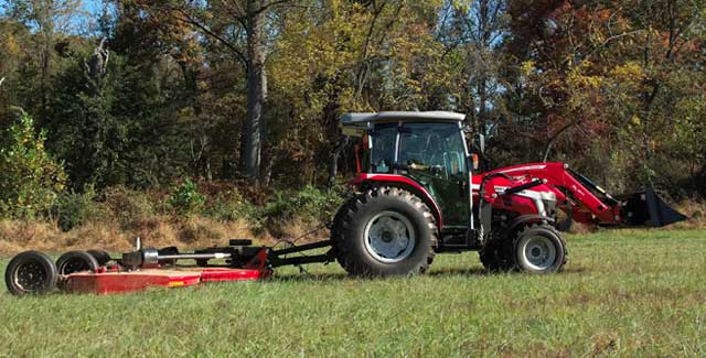A red Massey Ferguson 2M tractor pulling a large rotary mower across a grassy field bordered by tall trees in early autumn.