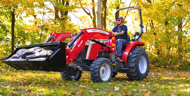 A person operates a red Massey Ferguson 1E Series compact tractor with a front loader full of mulch bags in a grassy, wooded area during fall, surrounded by colourful autumn leaves.