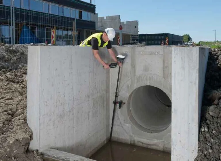 A construction worker in a hard hat and hi-vis vest leaning over a concrete culvert structure, using a Trimble GNSS survey rover on a pole to measure a large drainage pipe