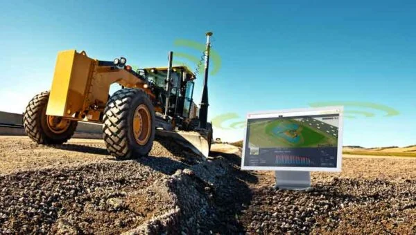 A yellow motor grader working on a gravel road with wireless signal icons and a monitor showing a 3D grade-control model, illustrating GPS machine guidance