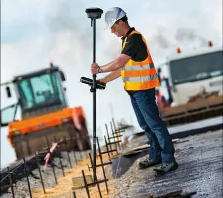 A surveyor in a hard hat and hi-vis vest holding a Trimble GNSS rover pole on a road construction site, with a roller and equipment in the background