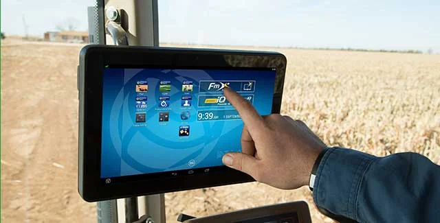 A hand tapping a Trimble FmX in-cab display screen mounted inside a tractor, with a field visible through the window