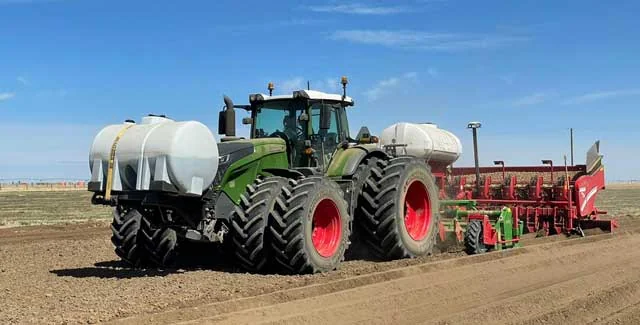 A green Fendt tractor pulling a planter with two white liquid tanks, working a freshly tilled field under a clear blue sky